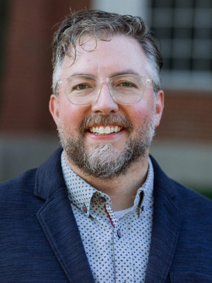 A man wearing glasses, a dark blue blazer, and a button up shirt stands outside smiling