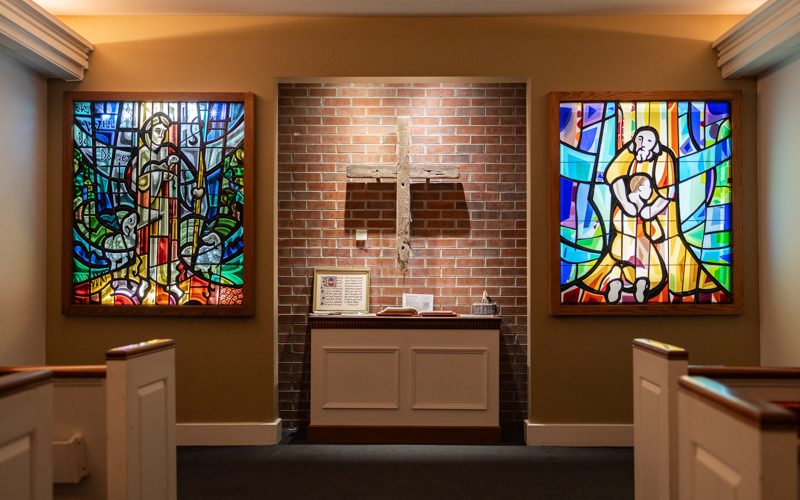 The front of chapel which contains a large tan cross hanging on brick wall in between two stain glass windows. In front of the cross lies a table containing a few items, including an open Bible.