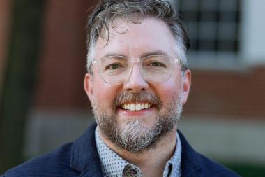 A man wearing glasses, a dark blue blazer, and a button up shirt stands outside smiling